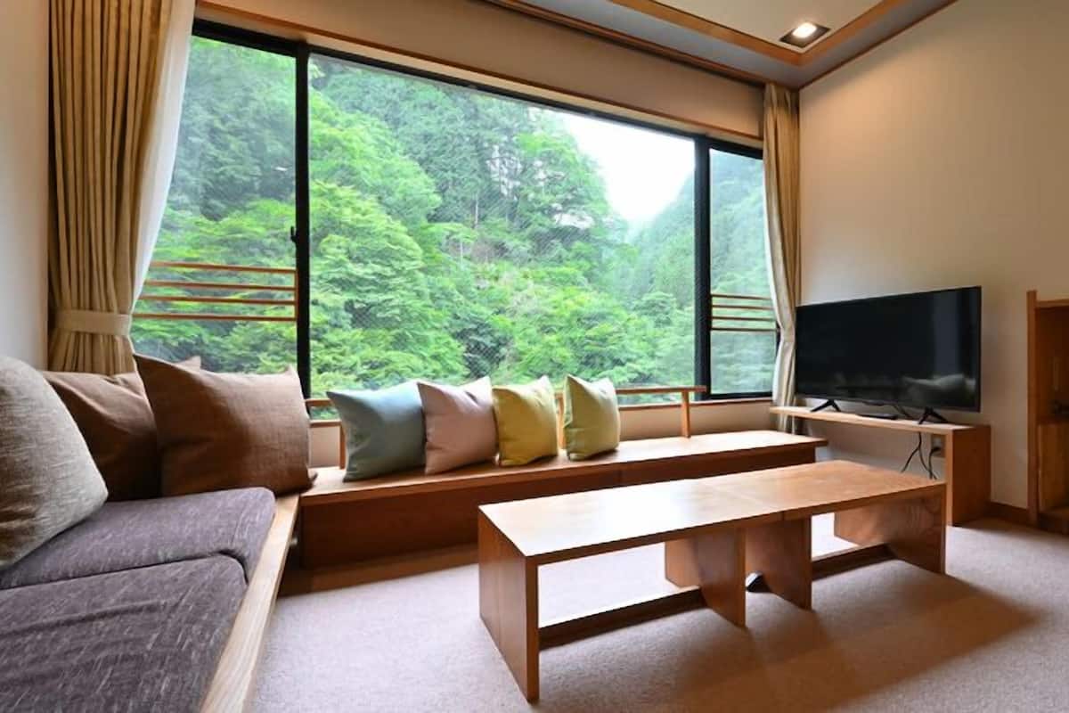 A living room with a sofa, a wooden coffee table, a TV on a stand, and a large window with a view of greenery.