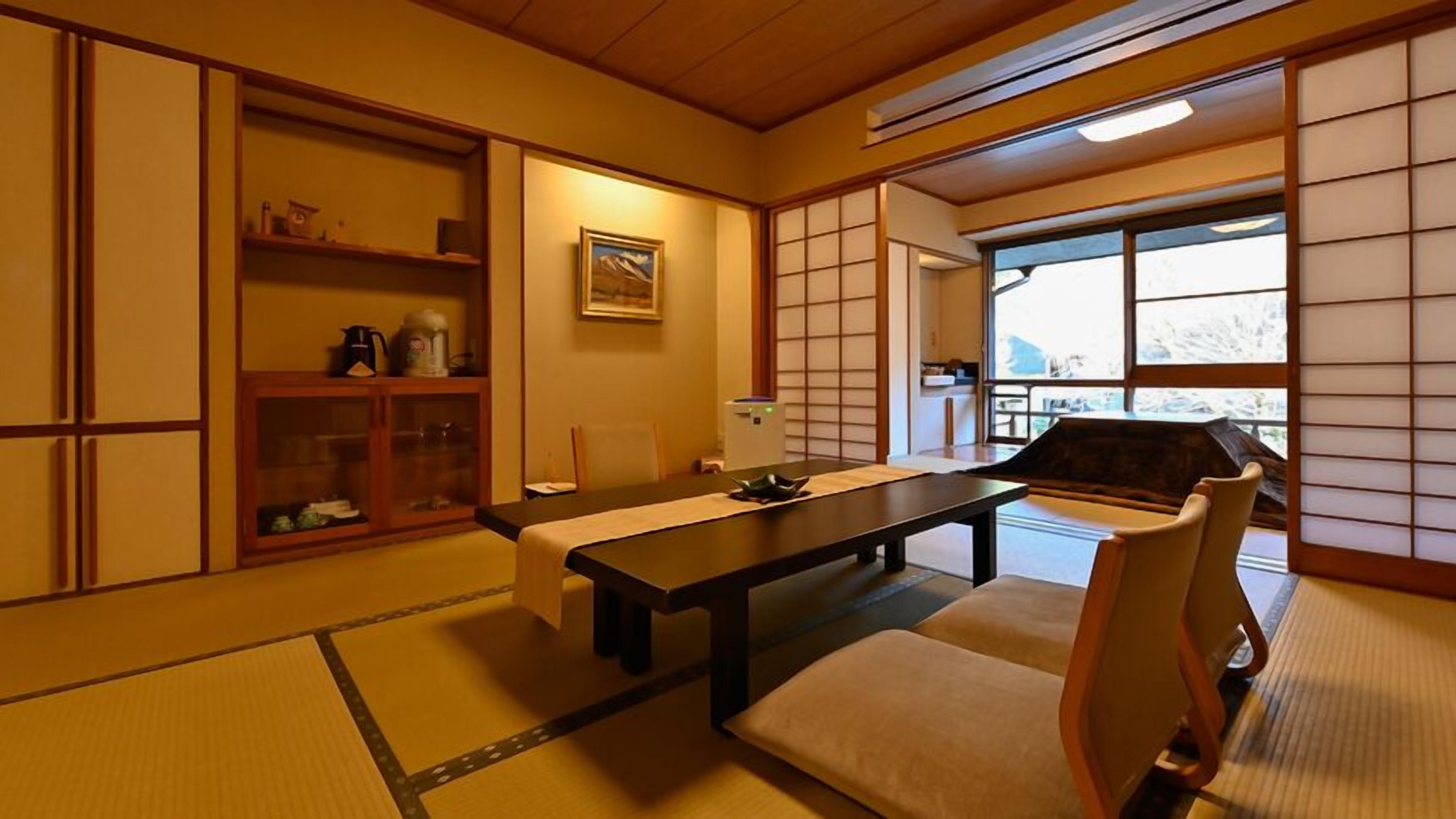 Ein traditioneller japanischer Raum mit Tatami-Boden, einem niedrigen Tisch mit Kissen, Schiebetüren und Blick durch ein Fenster.