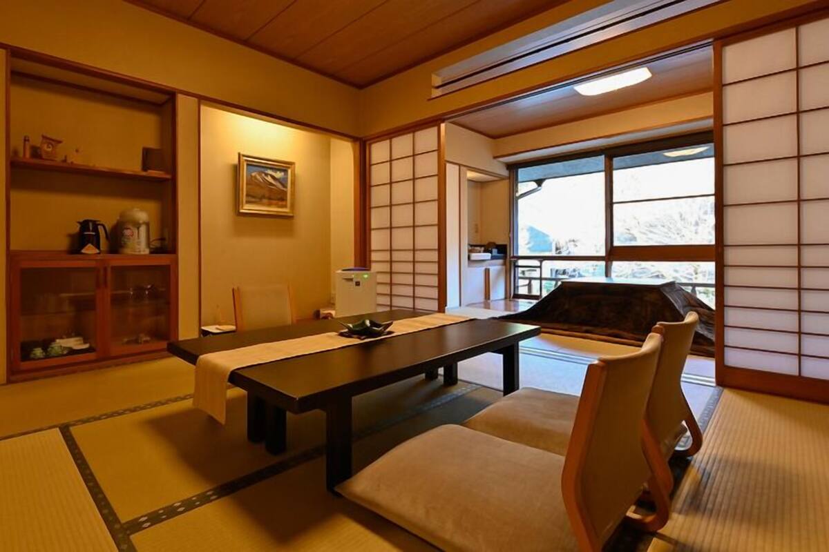 A traditional Japanese room with tatami flooring, a low table with cushions, sliding doors, and a view through a window.