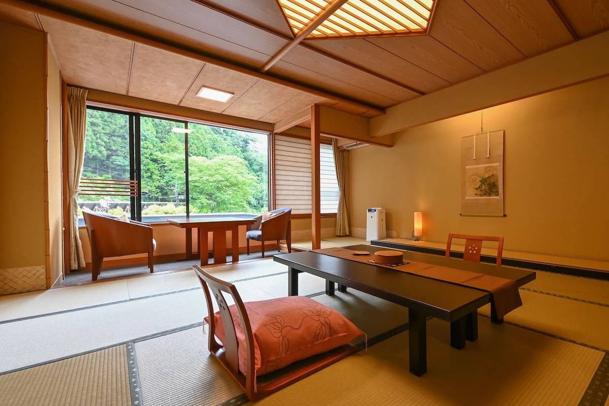 Une pièce de style japonais traditionnel, avec un sol recouvert de tatamis, une table basse, des chaises, une porte coulissante et une vue sur la verdure à l’extérieur.
