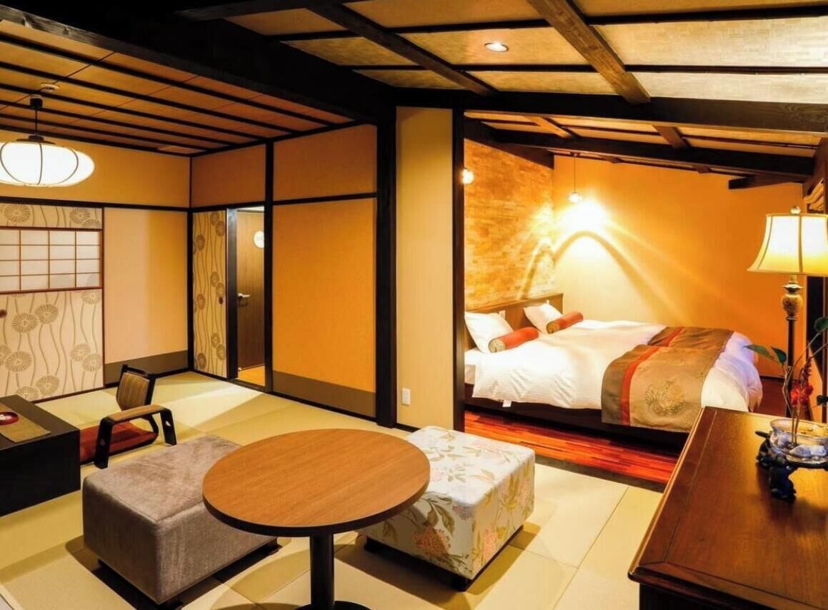 Habitación de hotel con suelo de tatami, una cama con edredón rojo y blanco, una mesa redonda de madera y una lámpara de estilo japonés tradicional.