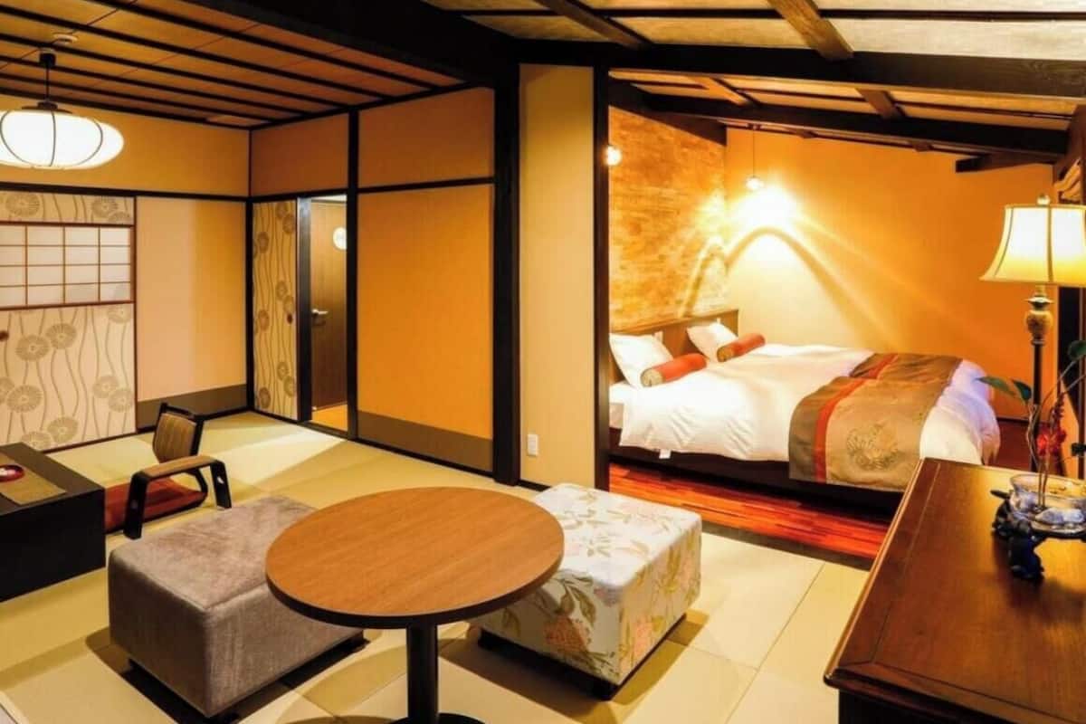 Habitación de hotel con suelo de tatami, una cama con edredón rojo y blanco, una mesa redonda de madera y una lámpara de estilo japonés tradicional.