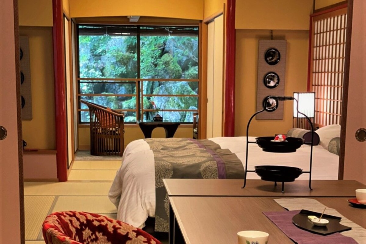 A traditional Japanese-style room with tatami flooring, a low table with a tea set, and a sliding door leading to a balcony with a view of trees.