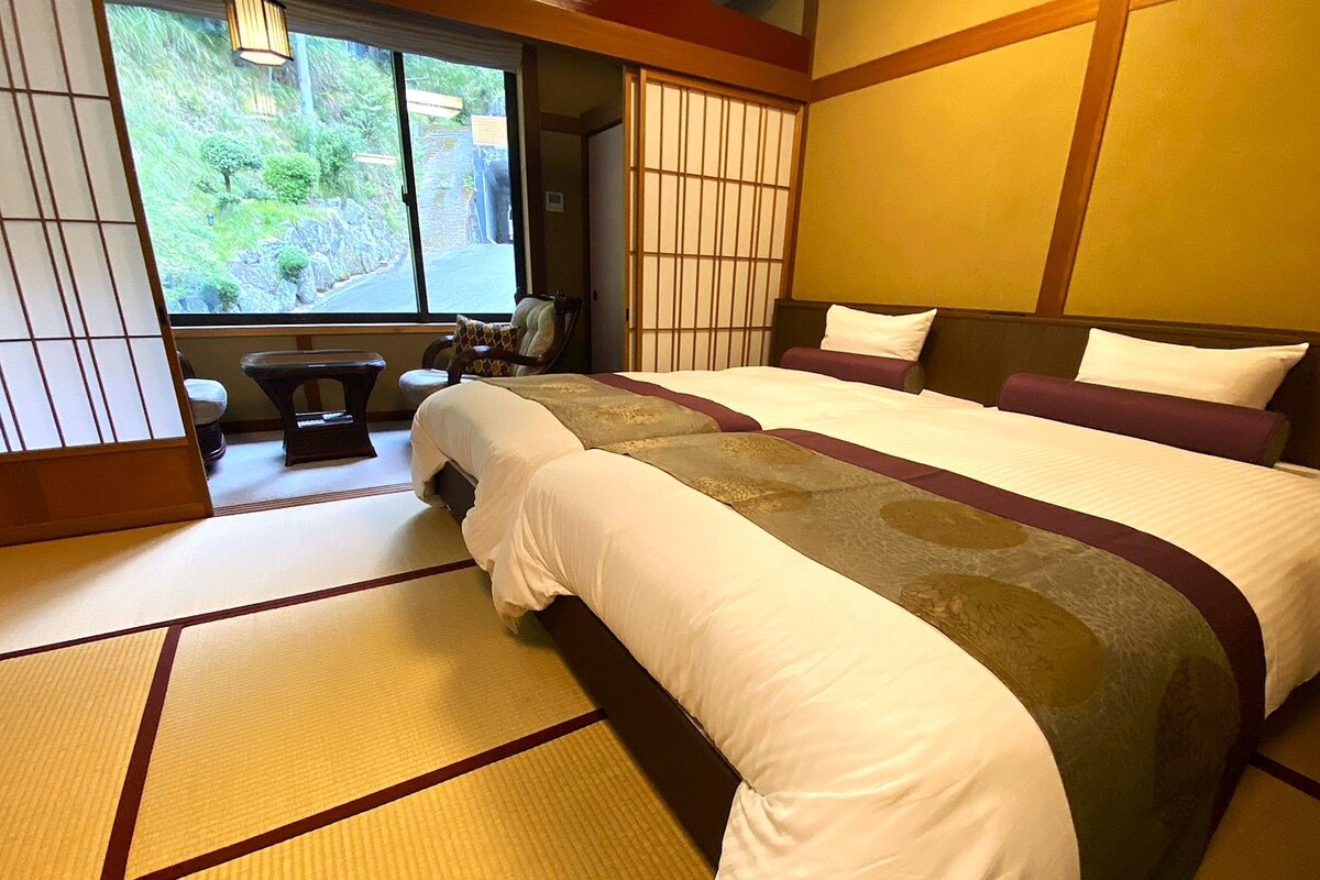 A traditional Japanese-style hotel room with tatami flooring, a large bed with white and brown bedding, a view of greenery outside, and a sliding door.