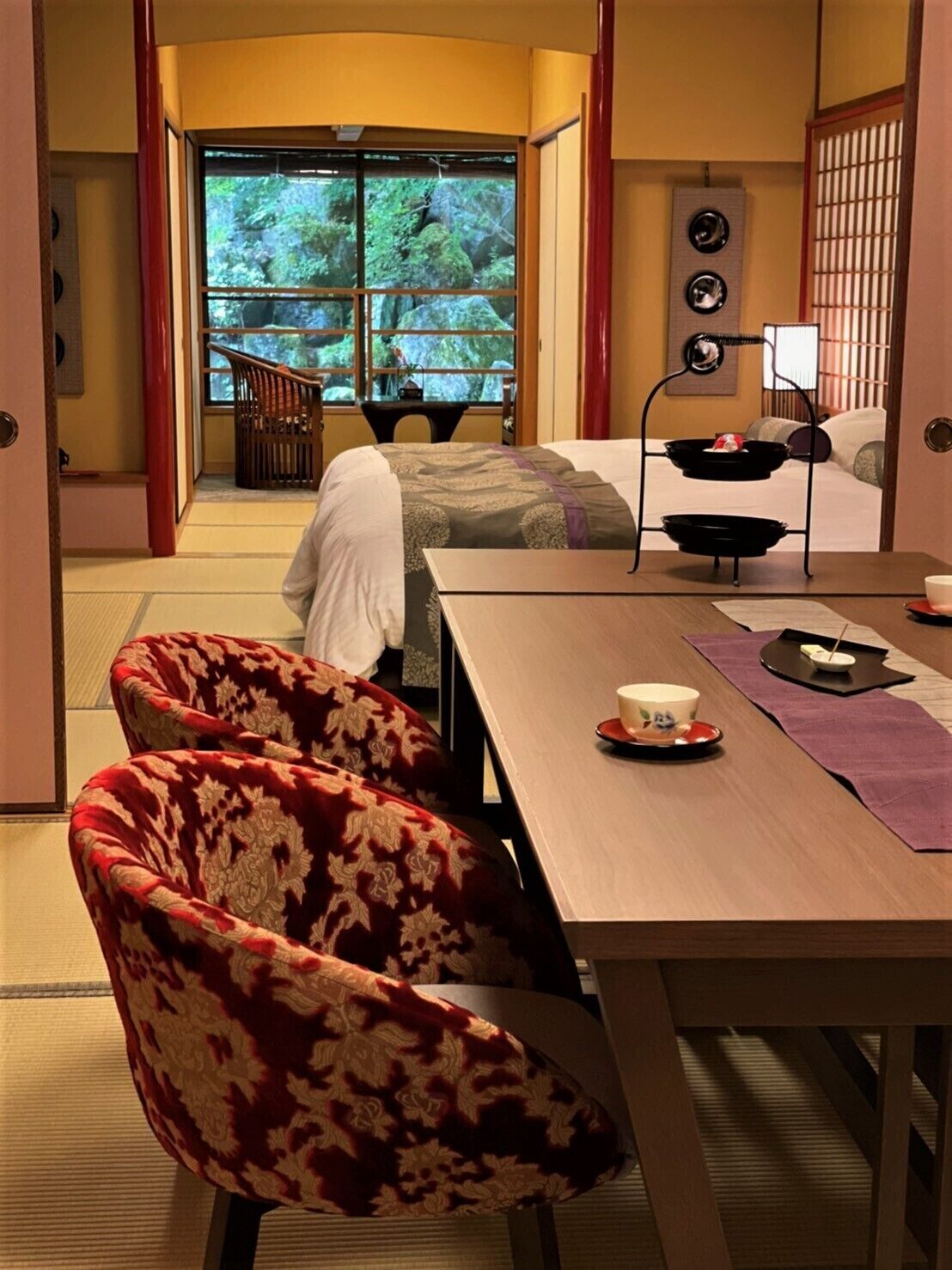 Una habitación tradicional japonesa con suelo de tatami, una mesa baja con un set de té y una puerta corrediza que da a un balcón con vistas a árboles.