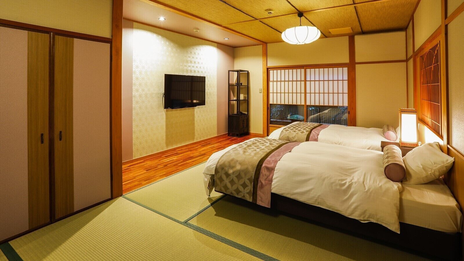 Una habitación tradicional japonesa con suelo de tatami, una cama con colcha estampada, un televisor montado en la pared y puertas correderas.