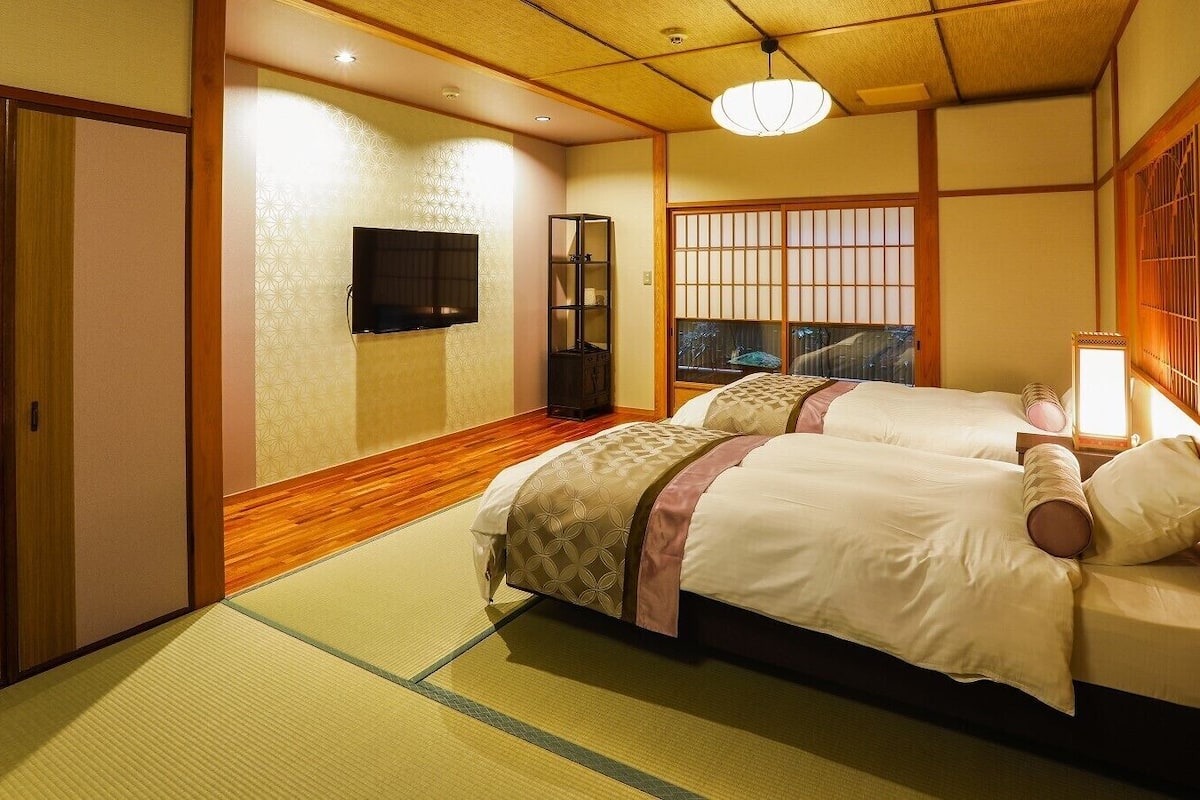 Una habitación tradicional japonesa con suelo de tatami, una cama con colcha estampada, un televisor montado en la pared y puertas correderas.