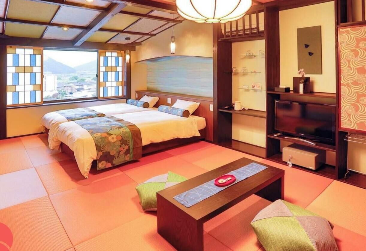 Habitación de hotel con esteras de tatami, una cama con cubrecama floral, televisión y vistas al exterior.