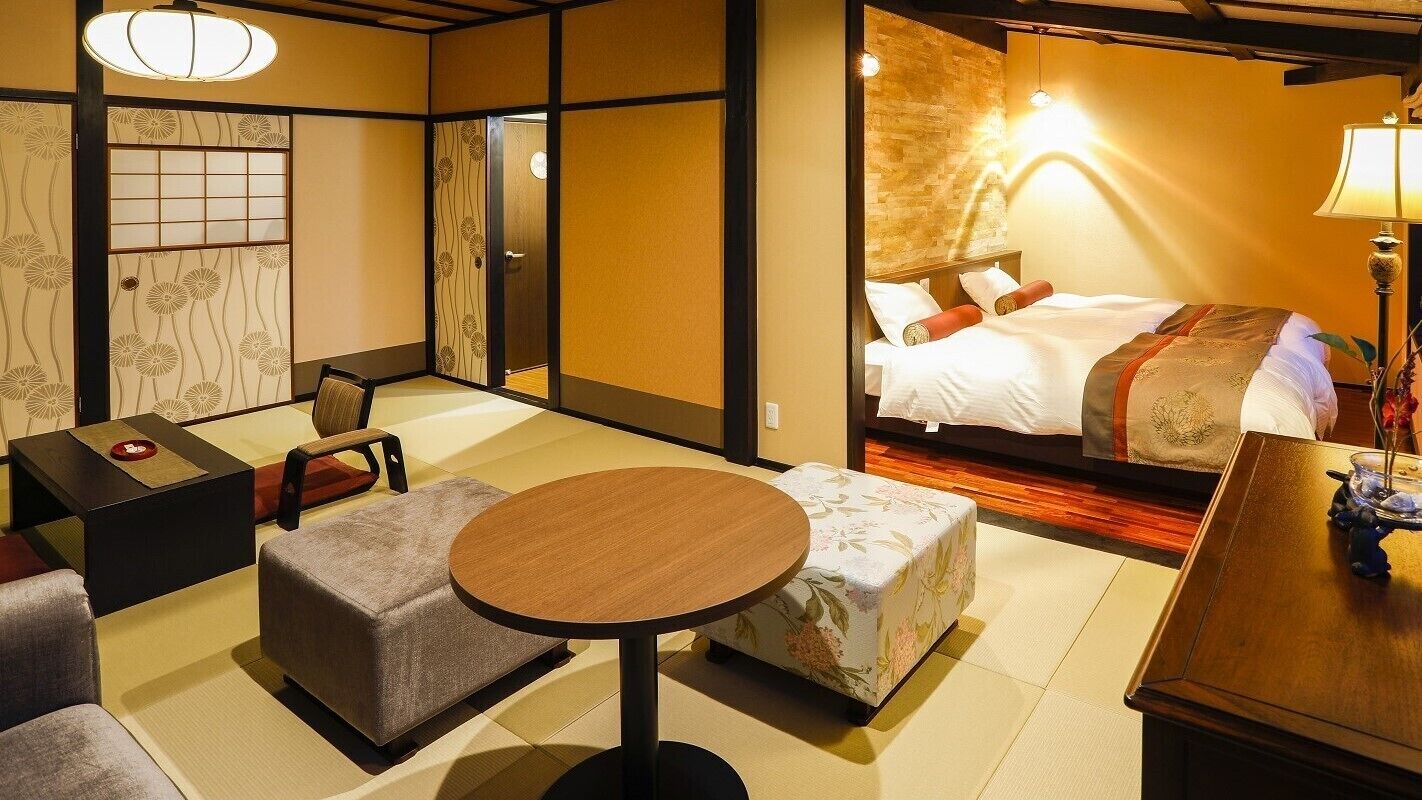 Habitación de hotel en estilo tradicional japonés, con suelo de tatami, una cama con cabecera de madera, una pequeña mesa redonda y un rincón de estar con cojines.