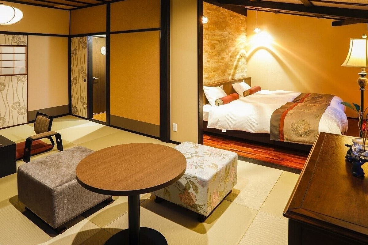 Habitación de hotel en estilo tradicional japonés, con suelo de tatami, una cama con cabecera de madera, una pequeña mesa redonda y un rincón de estar con cojines.