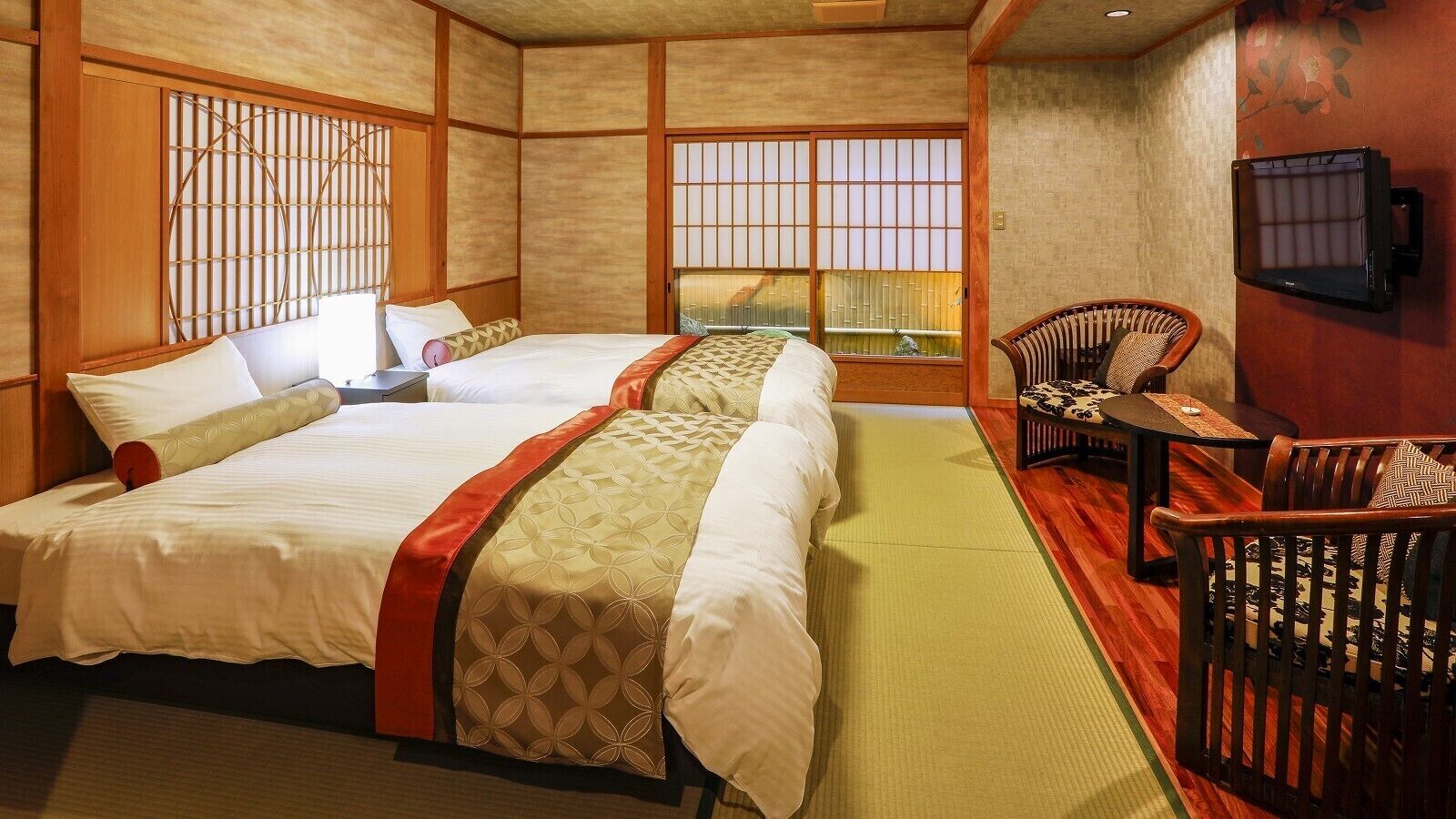 Una habitación de estilo japonés tradicional con suelo de tatami, dos camas con cubrecamas estampados, una mesa baja con una silla y un televisor montado en la pared.