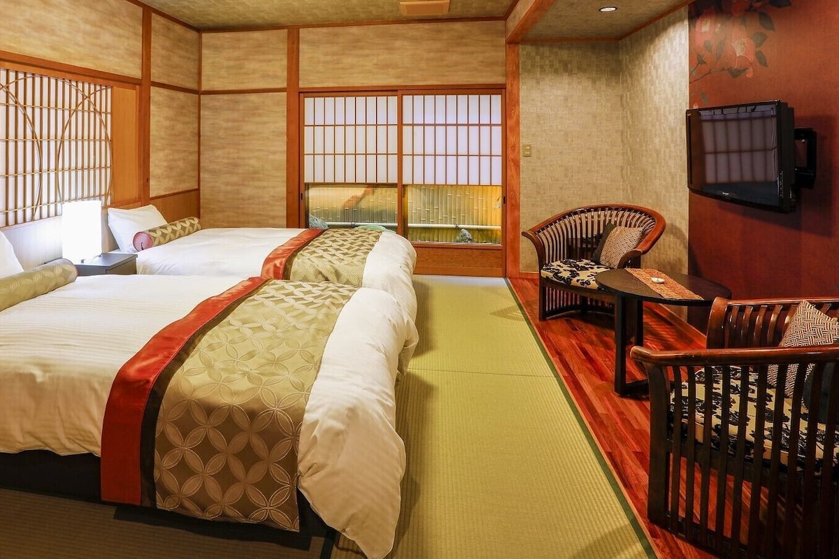 Una habitación de estilo japonés tradicional con suelo de tatami, dos camas con cubrecamas estampados, una mesa baja con una silla y un televisor montado en la pared.