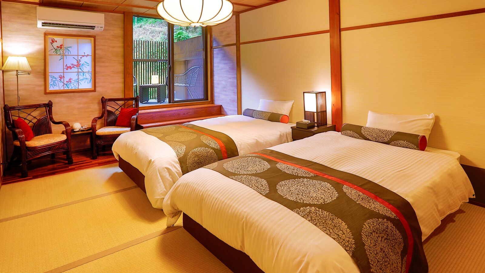 Una habitación japonesa tradicional con suelo de tatami, dos camas con ropa de cama estampada, un escritorio de madera, sillas y un fusuma que da a un área exterior.