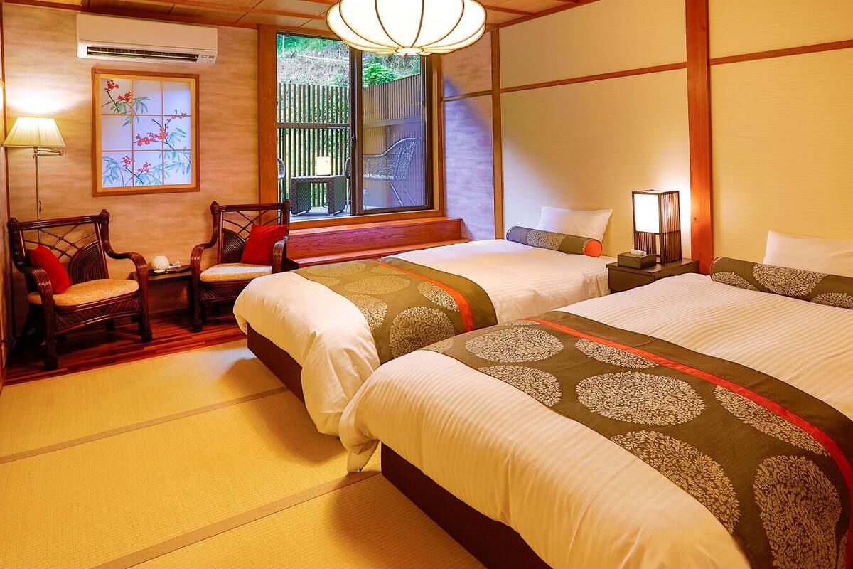 Ein traditioneller japanischer Raum mit Tatami-Boden, zwei Betten mit gemusterten Bettwäsche, einem Holztisch, Stühlen und einer Schiebetür zu einem Außenbereich.