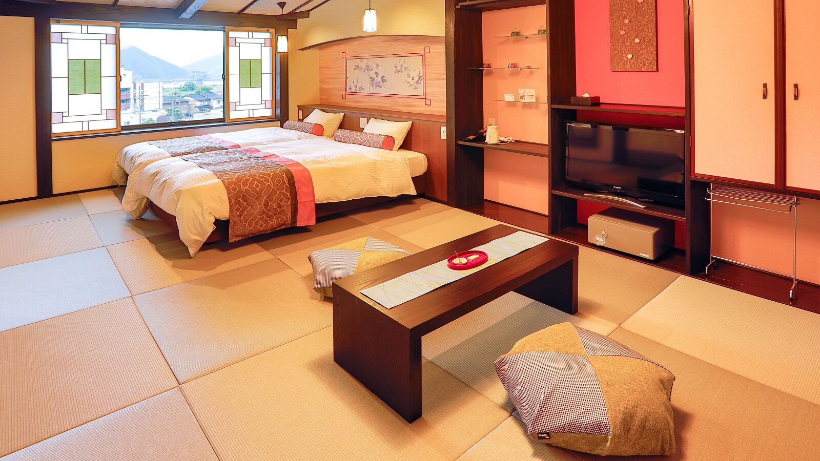 Una habitación tradicional japonesa con suelo de tatami, una cama con manta de estampado rojo y blanco, una mesa de madera con un objeto rojo, y un televisor sobre un mueble.