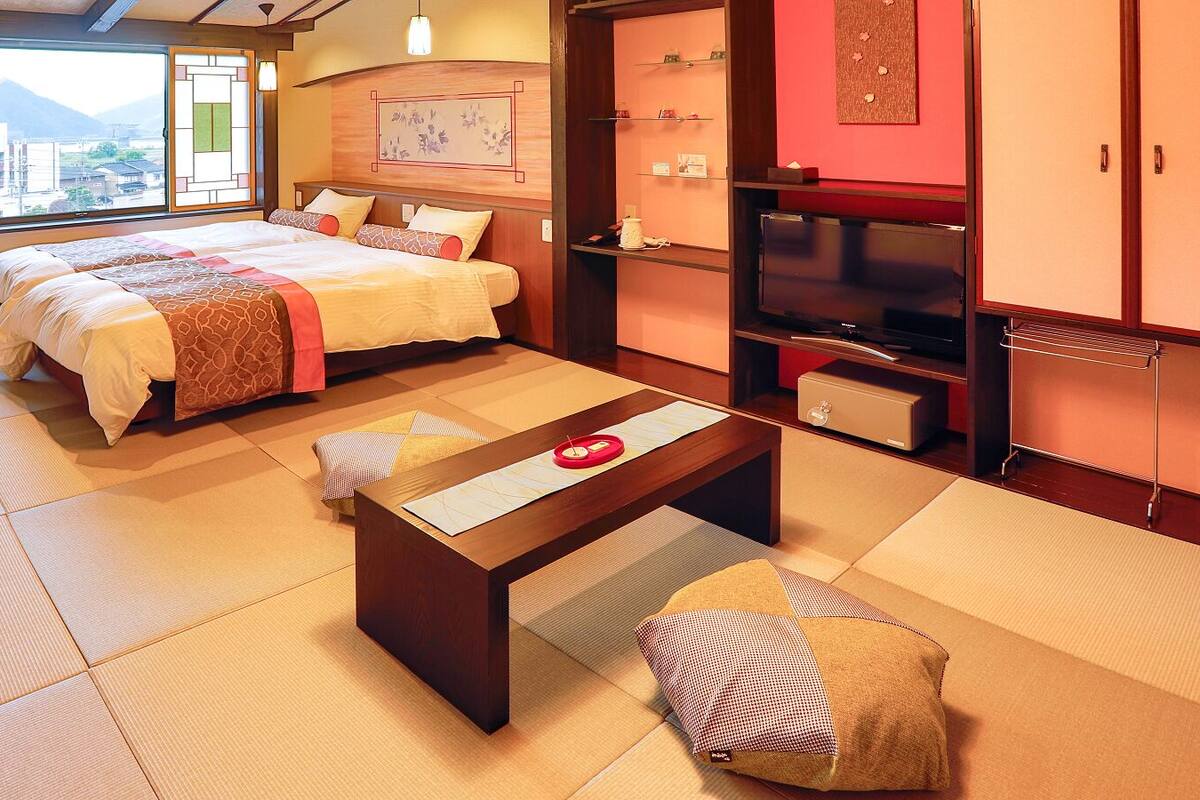 Ein traditionell eingerichteter japanischer Raum mit Tatami-Böden, einem Bett mit einer Decke in Rot-Weiß-Muster, einem Holztisch mit einem roten Gegenstand und einem Fernseher auf einem Standfuß.
