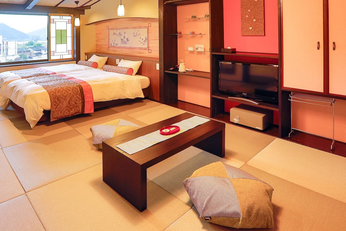 Una habitación tradicional japonesa con suelo de tatami, una cama con manta de estampado rojo y blanco, una mesa de madera con un objeto rojo, y un televisor sobre un mueble.