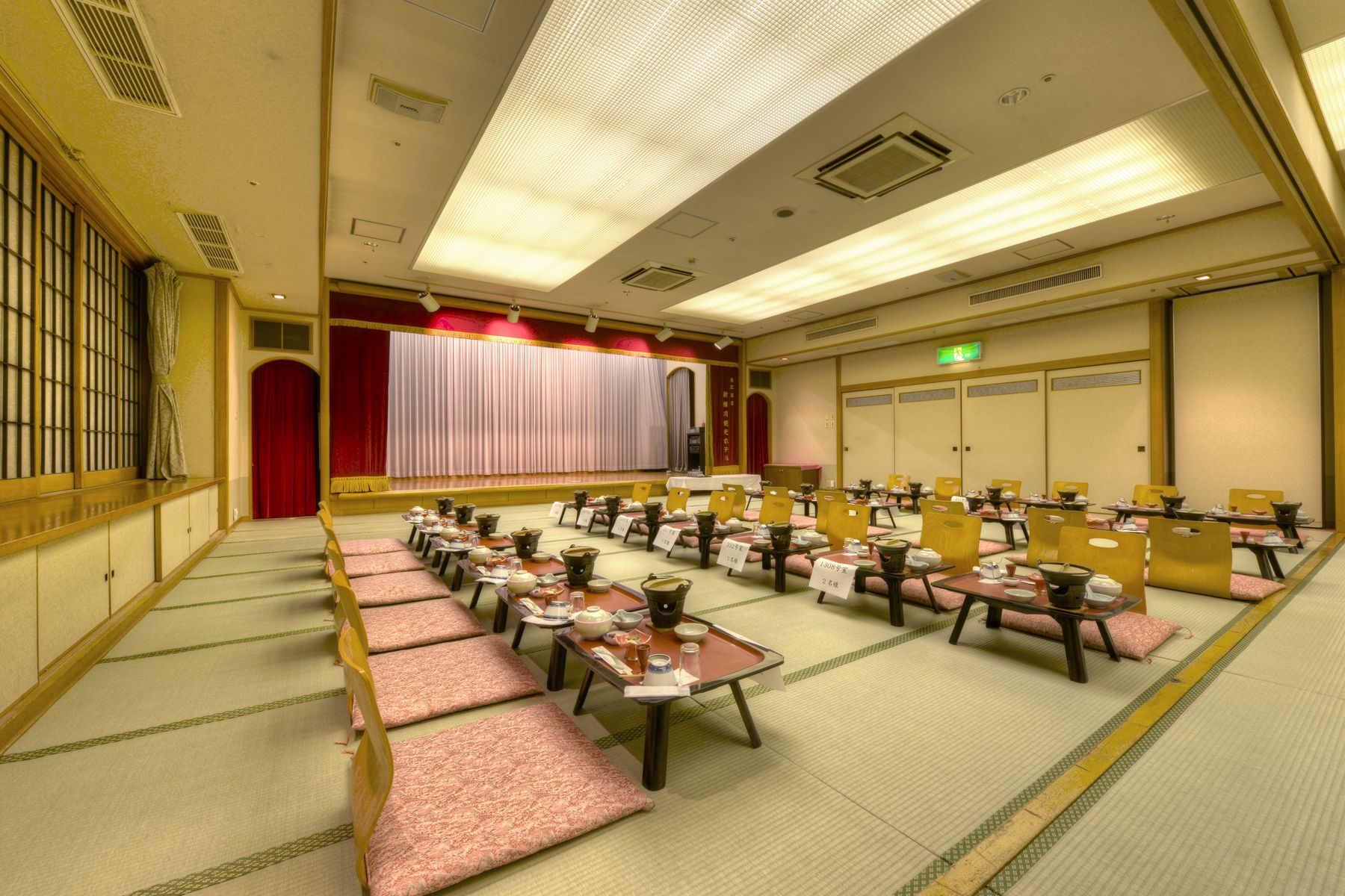 A conference room with rows of tables and chairs, a stage with curtains, and a traditional Japanese tatami mat floor.