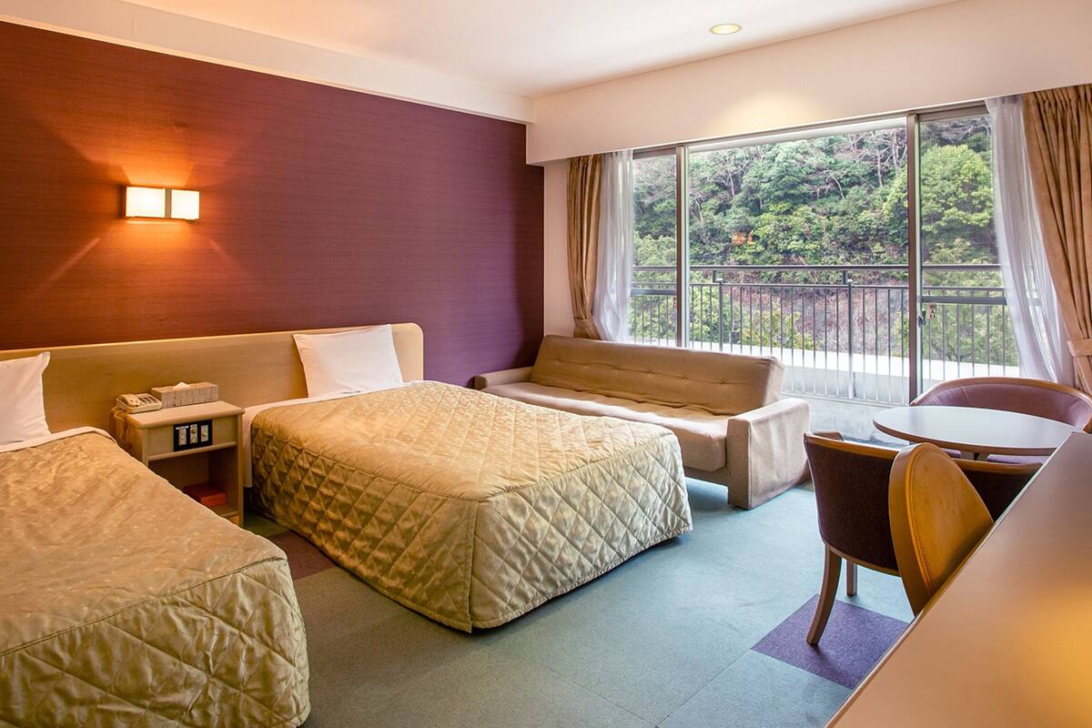 Een hotelkamer met twee bedden, een bank, een tafeltje en een stoel. Er is een raam met uitzicht op groen.