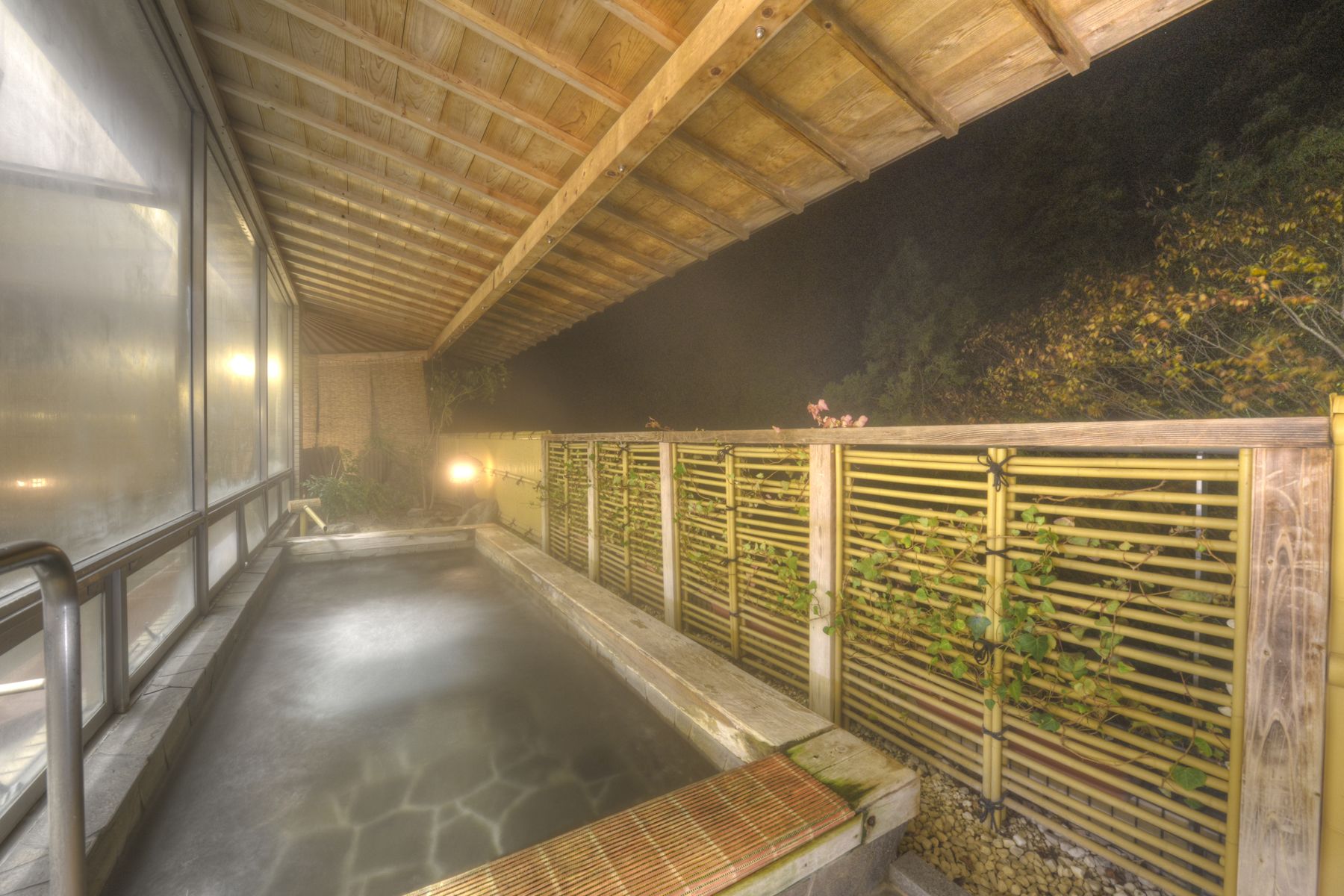 Un espace de baignade dans une source thermale, doté d’un plafond en bois, d’un sol en pierre et de rampes en bambou.