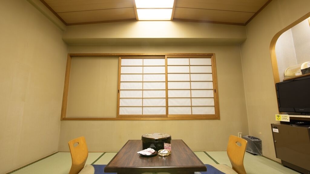 Une pièce avec une table recouverte d’un tatami, deux chaises en bois, une petite table avec un plateau, une fenêtre dotée de panneaux coulissants japonais (shoji) et un téléviseur posé sur un meuble.