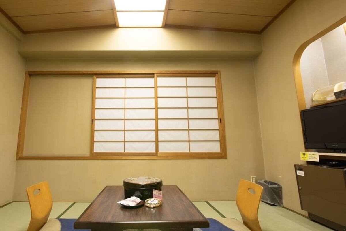 Une pièce avec une table recouverte d’un tatami, deux chaises en bois, une petite table avec un plateau, une fenêtre dotée de panneaux coulissants japonais (shoji) et un téléviseur posé sur un meuble.