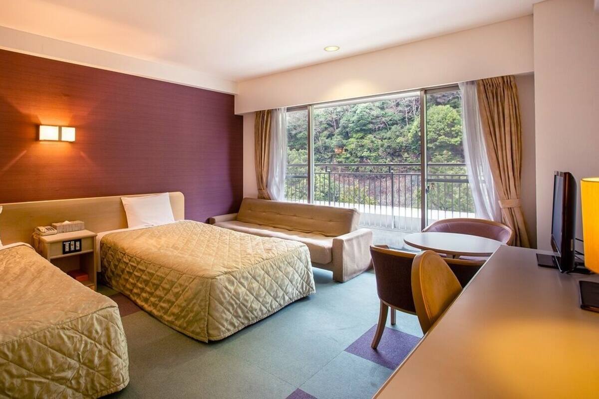 Hotelkamer met twee bedden, een bank, een bureau en een balkon met uitzicht op groen.