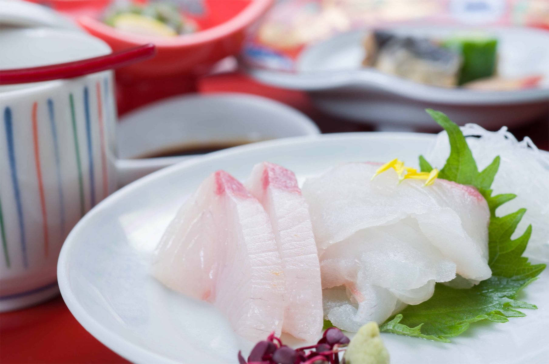 A plate of sashimi with a garnish of green leaves and yellow flowers, accompanied by a small bowl of soy sauce.