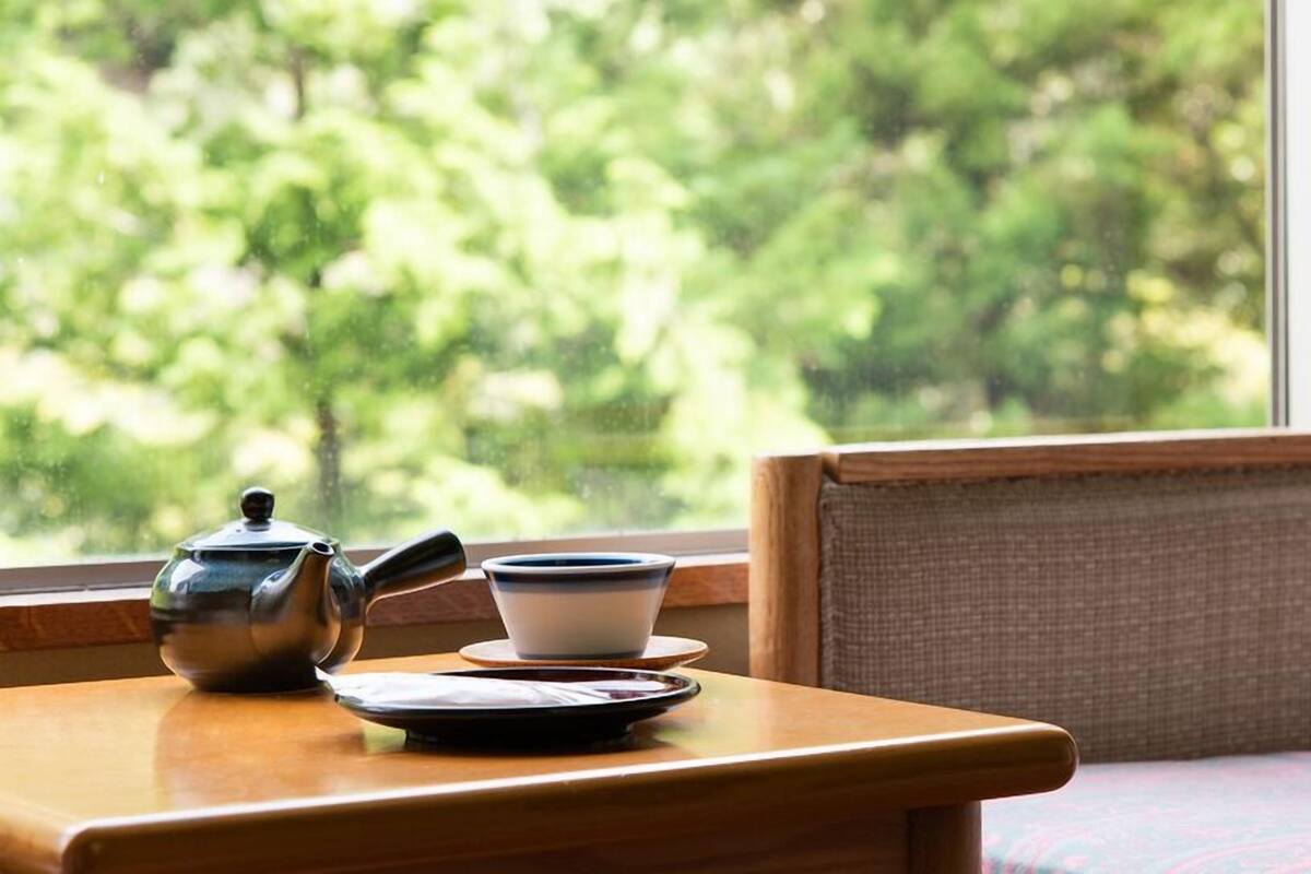 Een houten tafel met een theepot en kopjes, een raam met uitzicht op bomen, en een rieten stoel.