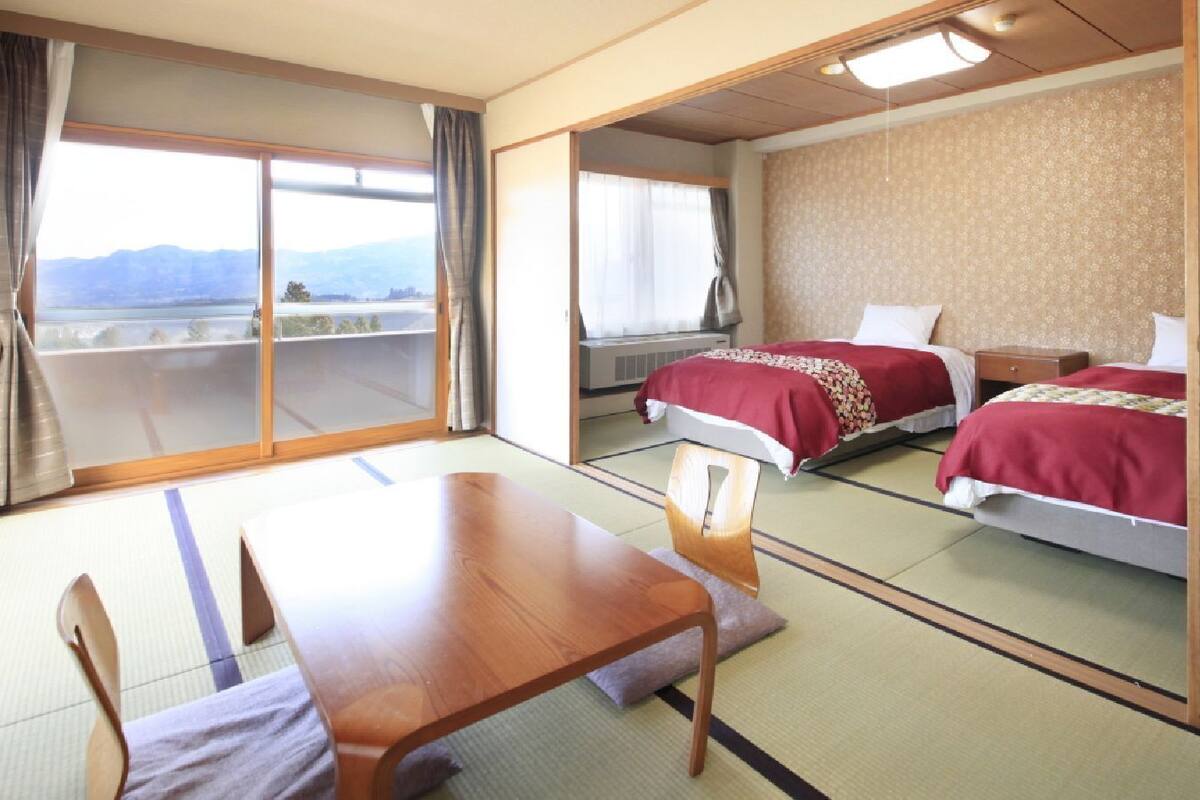 A hotel room with a wooden table, two beds with red bedding, a large window with a view, and tatami flooring.