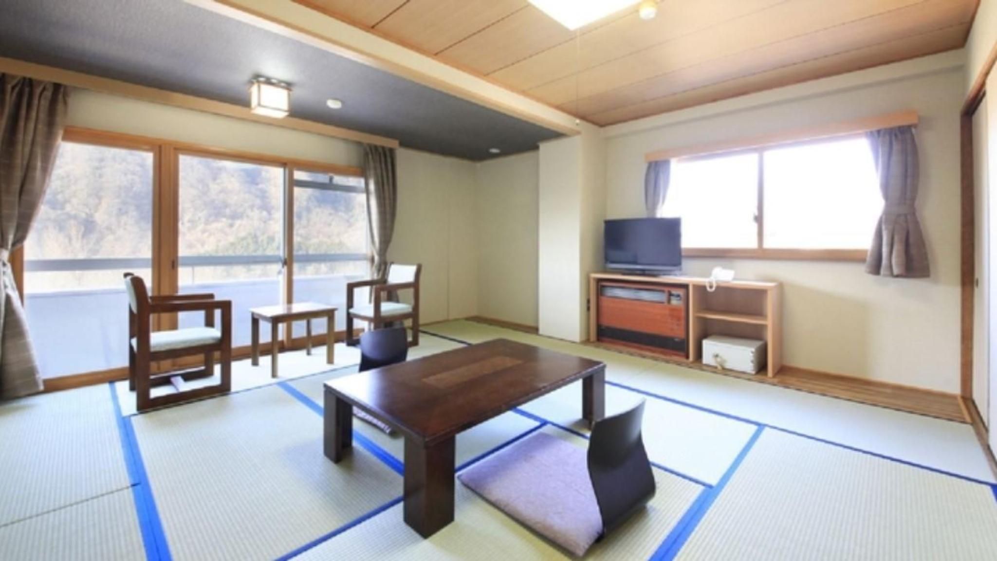 Ein traditioneller japanischer Raum mit Tatami-Bodenbelag, einem niedrigen Tisch, Stühlen, einem Fernseher und einer Schiebetür, die auf einen Balkon führt.
