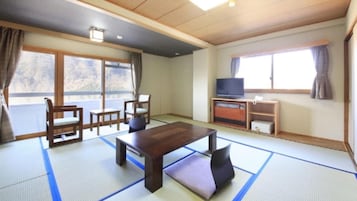 Una habitación tradicional japonesa con suelo de tatami, una mesa baja, sillas, un televisor y una puerta corredera que da a un balcón.