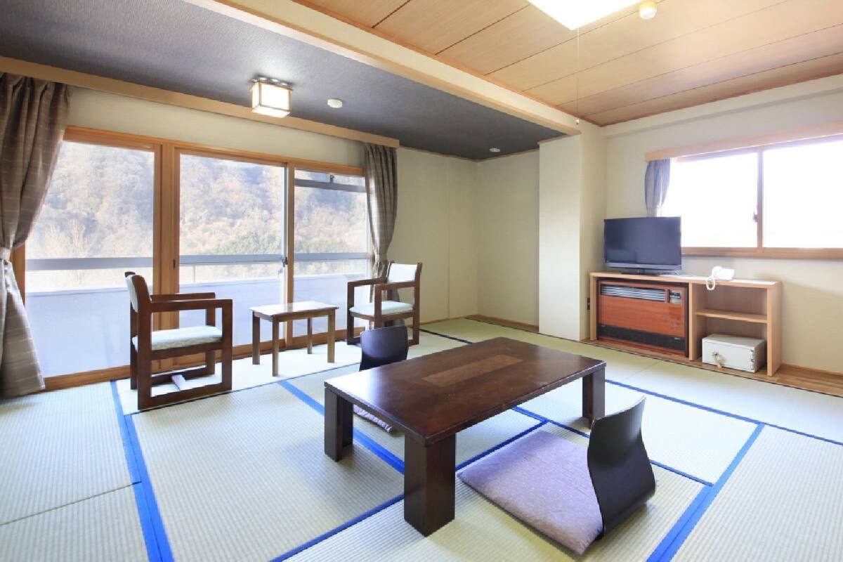 A traditional Japanese room with tatami flooring, a low table, chairs, a television, and a sliding door leading to a balcony.