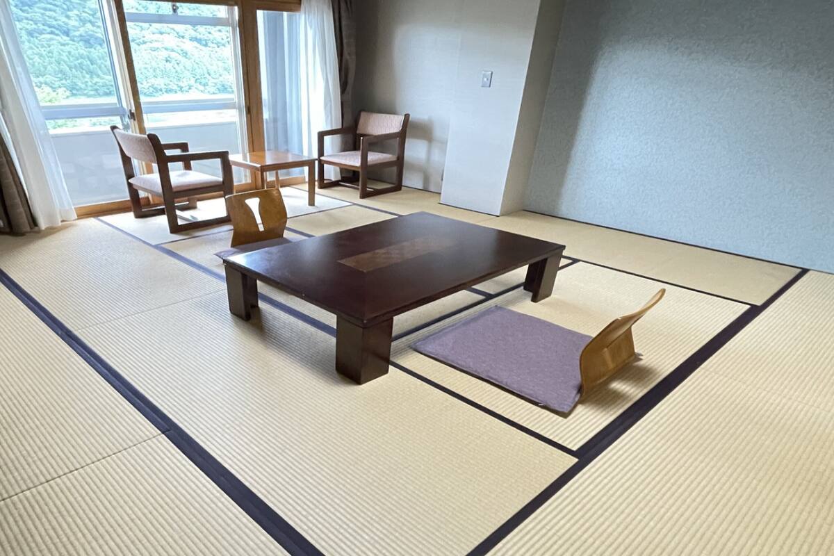 A tatami mat room with a low table, two chairs, and a view of a forest through large windows.