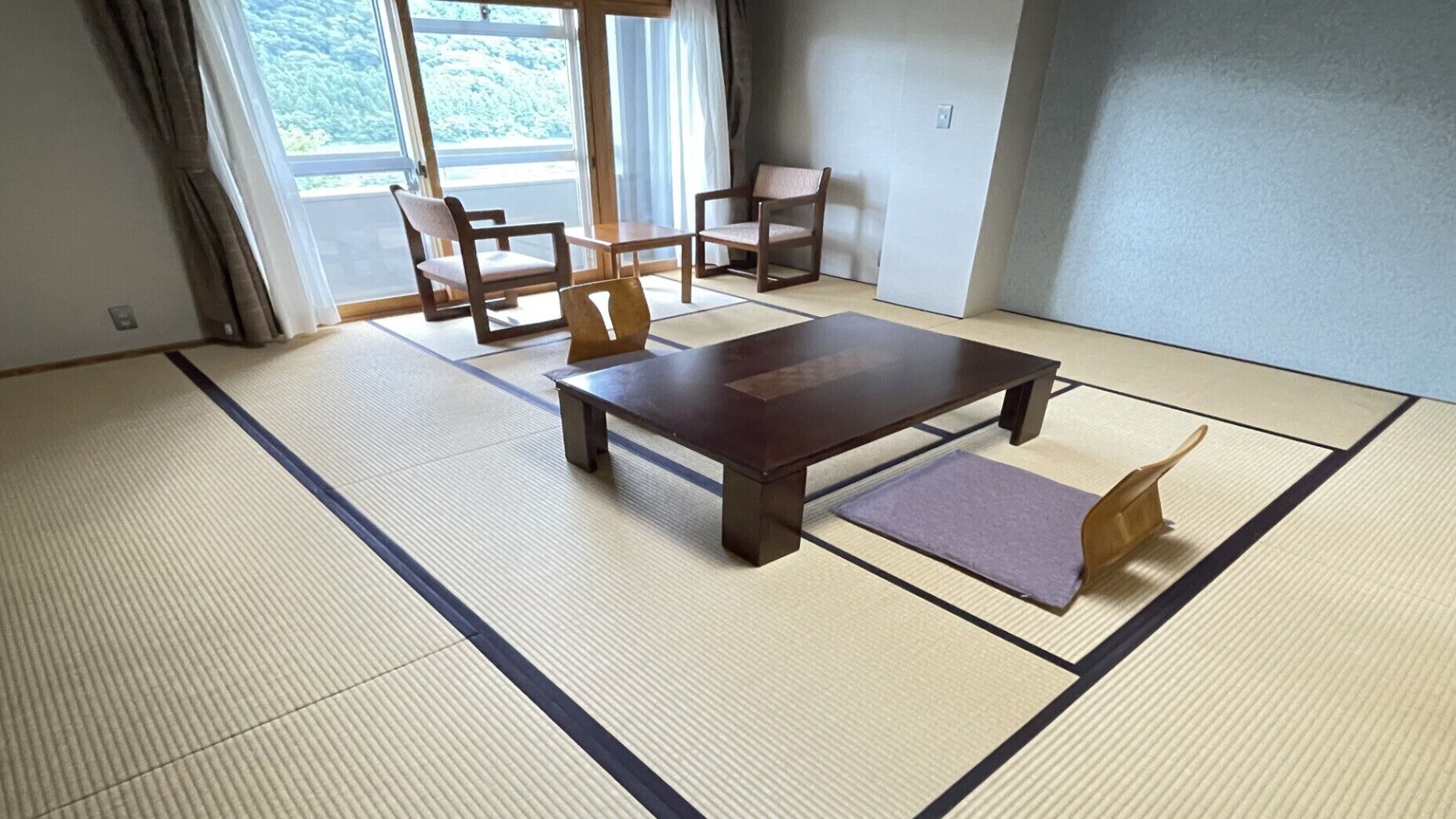 A tatami mat room with a low table and two chairs, large windows with curtains, and a view of greenery outside.