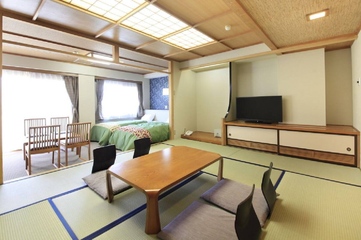 A traditional Japanese-style room with tatami flooring, a low table, chairs, a bed, and a television.