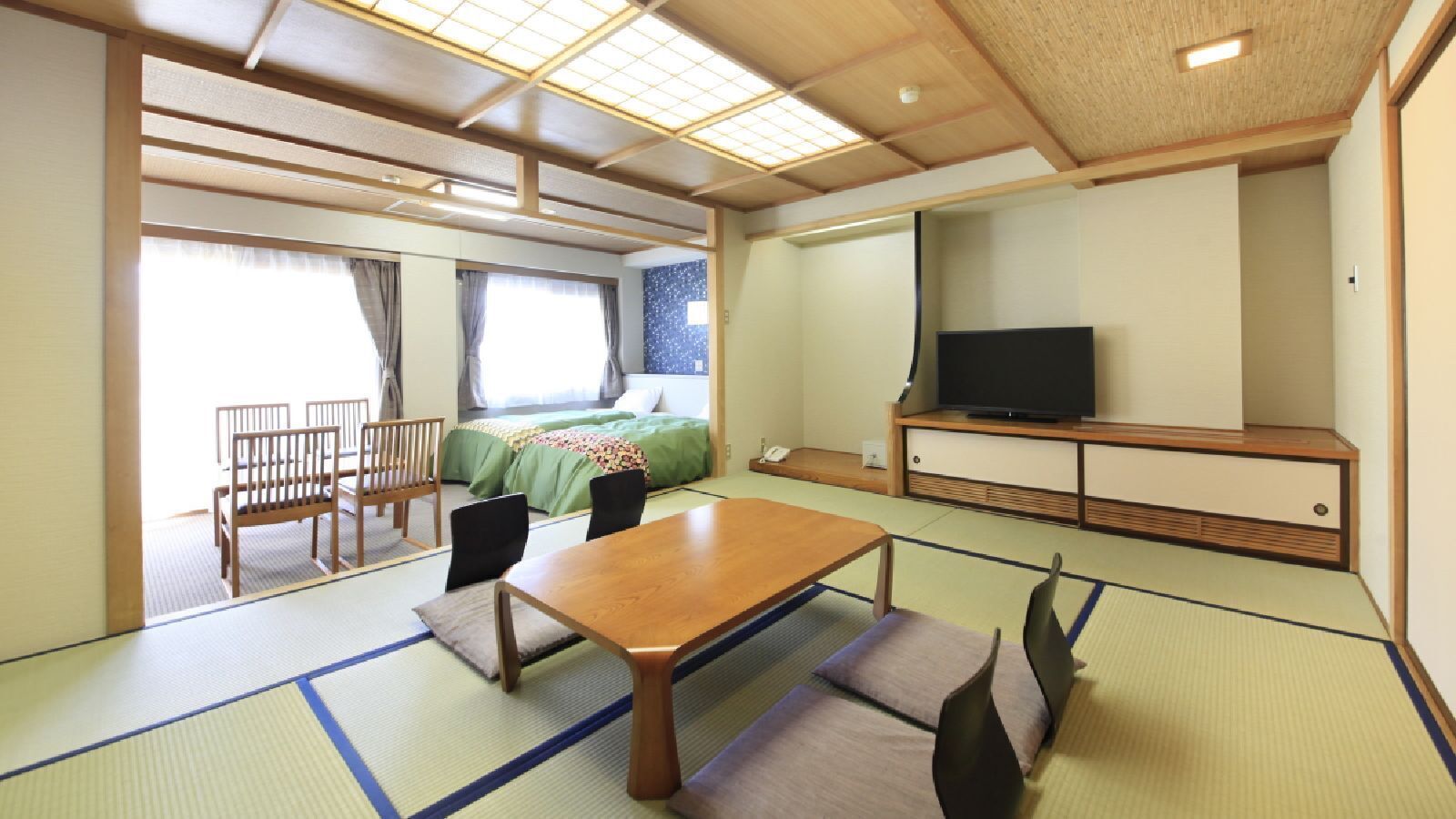 Habitació japonesa tradicional amb fosa de tatami, una taula baixa, cadires, un llit amb roba de llit verda, una televisió i una porta corredissa.
