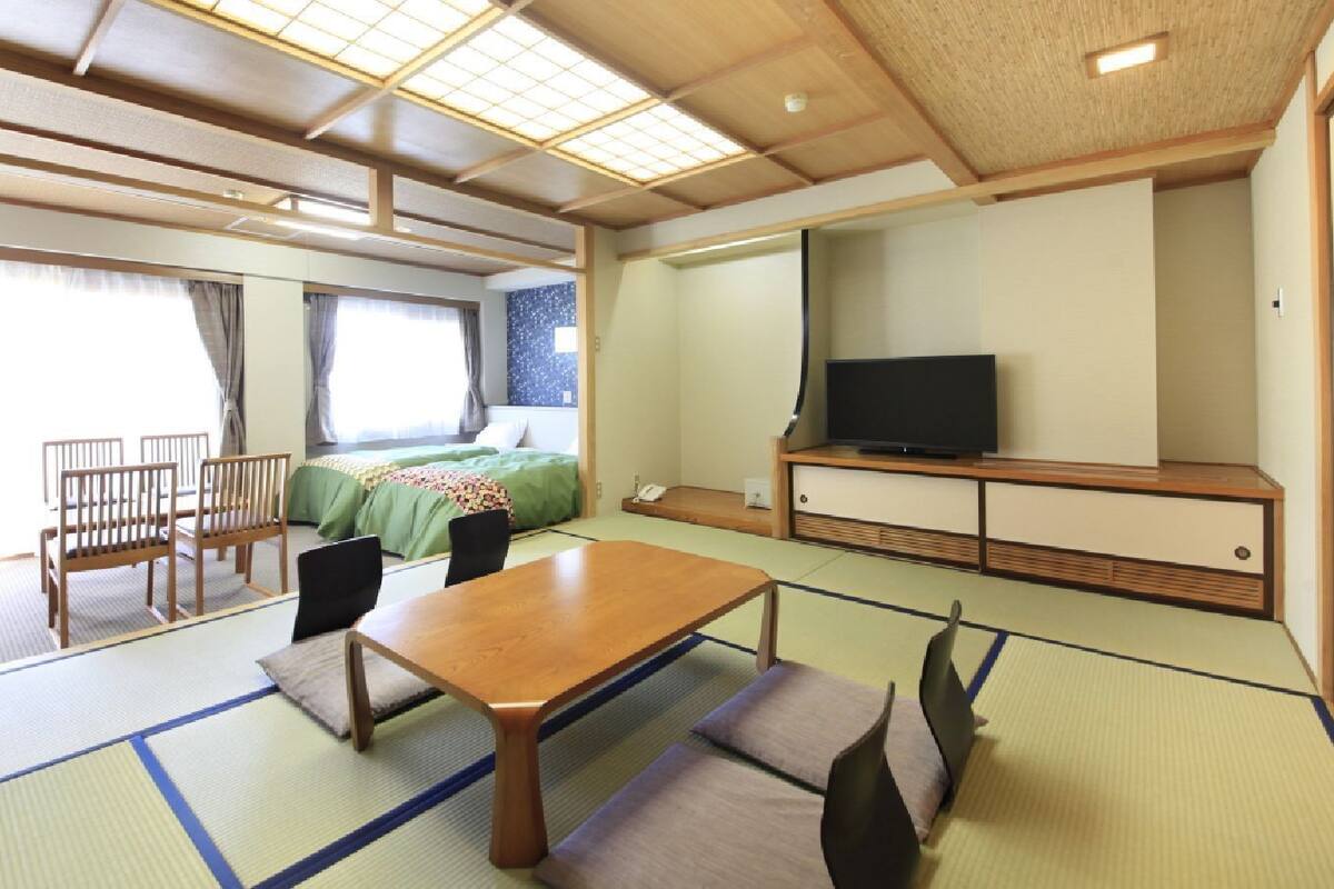 A traditional Japanese room with tatami flooring, a low table, chairs, a bed with green bedding, a television, and a sliding door.
