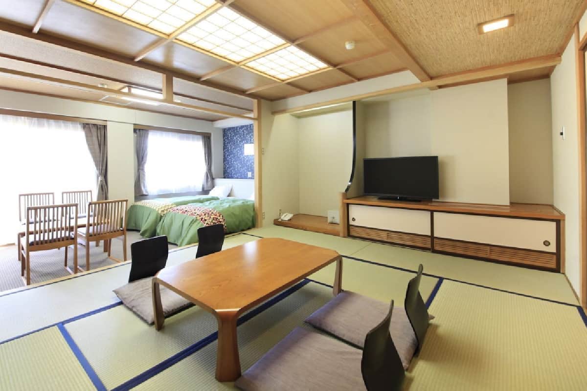 A traditional Japanese room with tatami flooring, a low table, chairs, a bed with green bedding, a television, and a sliding door.