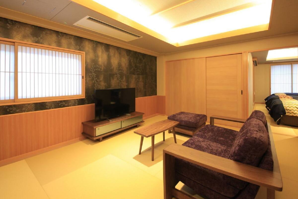 A living room with tatami flooring, a television on a low stand, a sofa, a small table, and sliding doors leading to a bedroom.