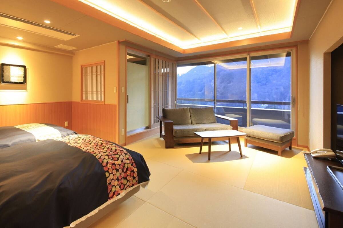 A hotel room with a bed, a sofa, a small table, a TV, and a view of mountains through large windows.