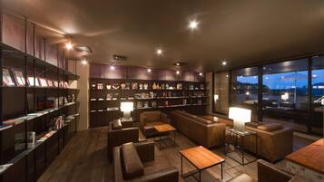 A modern lounge area with leather seating, a bookshelf, and a large window offering a view of the cityscape at dusk.