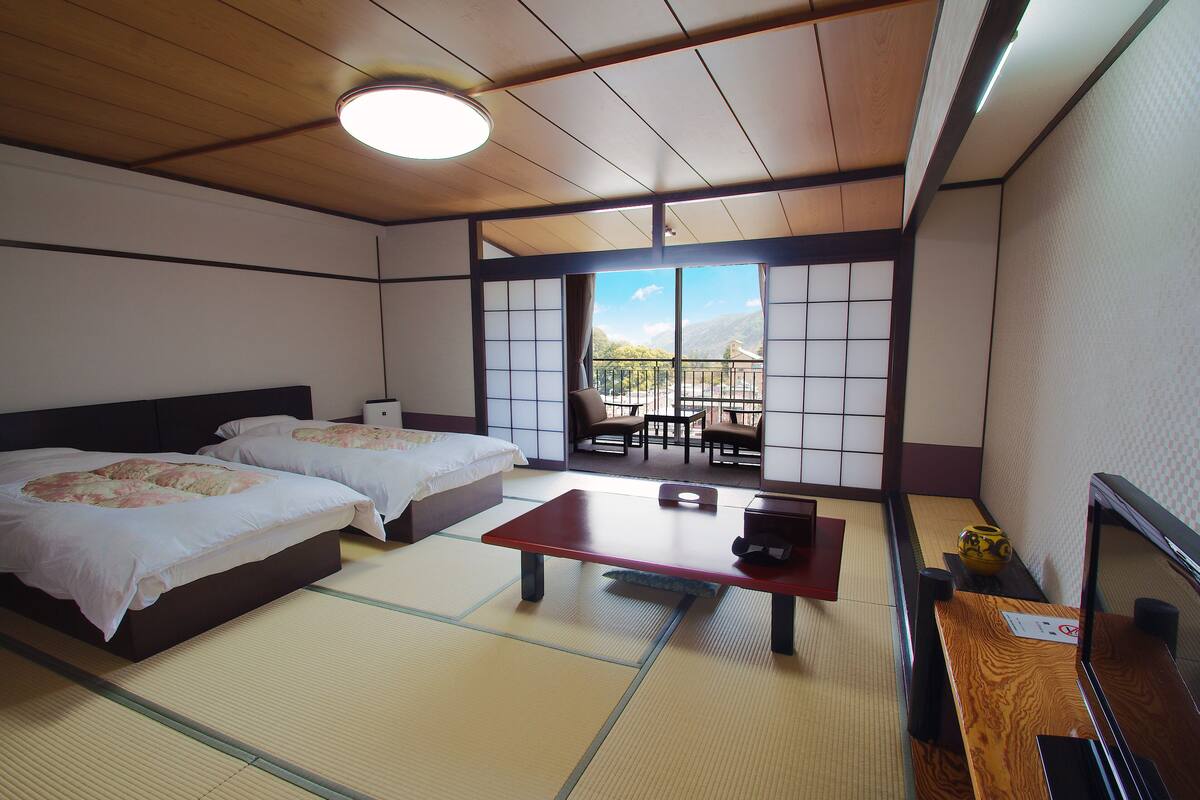 Una habitación japonesa tradicional con suelo de tatami, dos camas, una mesa de madera y vistas al exterior.