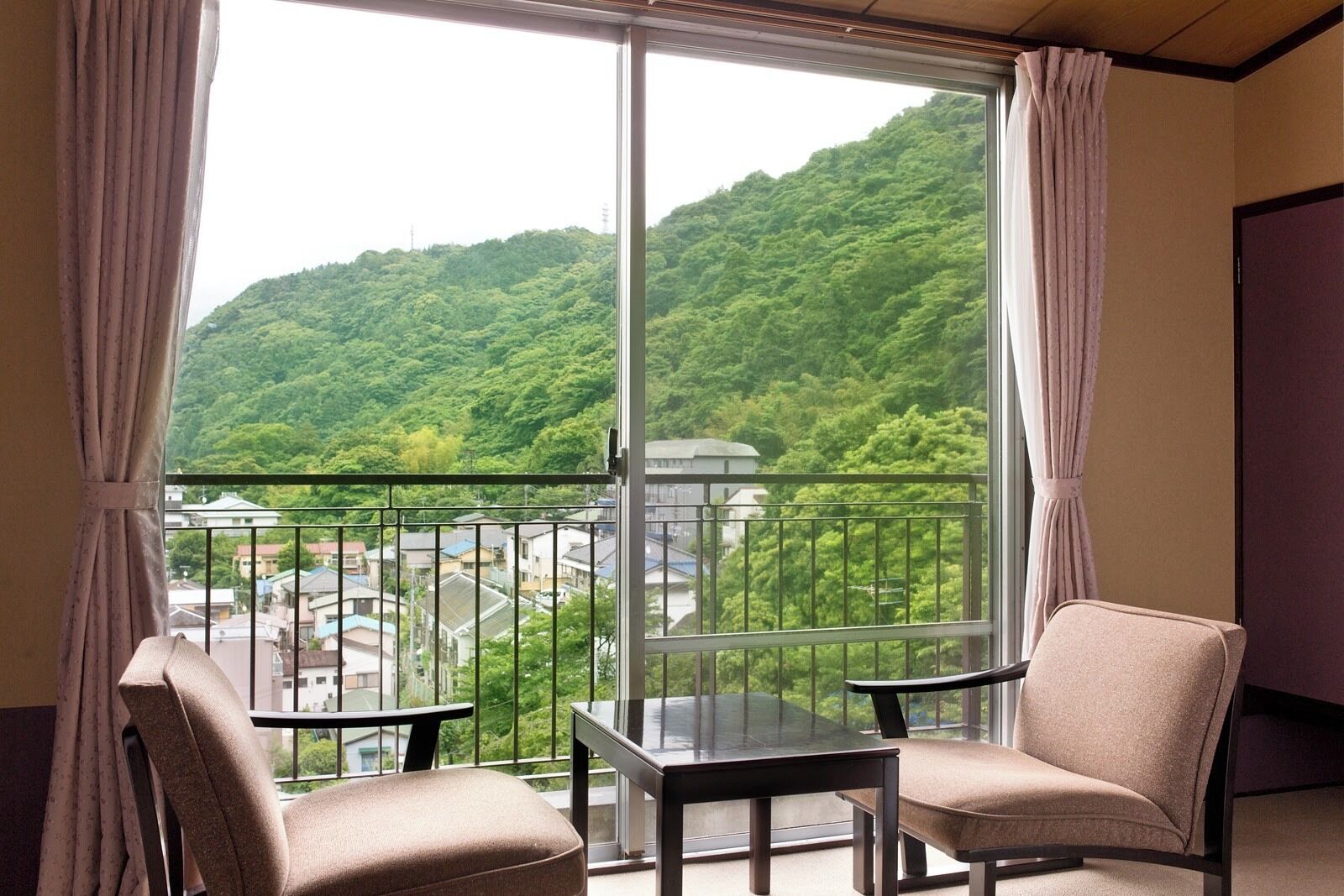 A balcony with two chairs and a table, a view of a green hillside, and a building in the background.