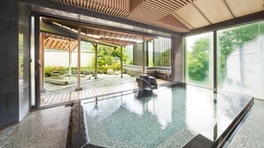 Piscina interior moderna con techo de madera y paredes de vidrio, que ofrece vistas a un jardín y un área de estar al aire libre.