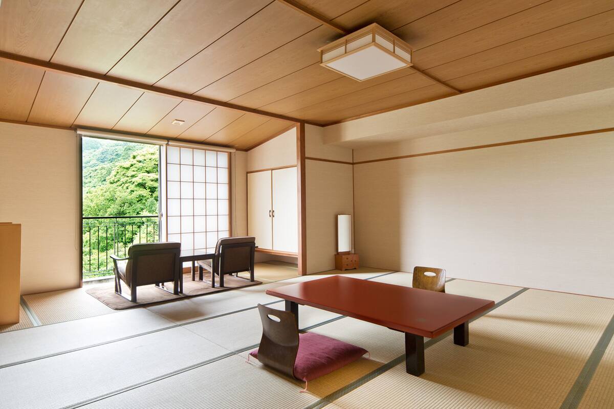 Una habitación japonesa tradicional con suelo de tatami, una mesa baja, dos sillas y una puerta corredera que da a un balcón con vistas a zonas verdes.