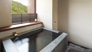Un baño moderno con una bañera grande, una ventana con persianas y vistas a una ladera verde.