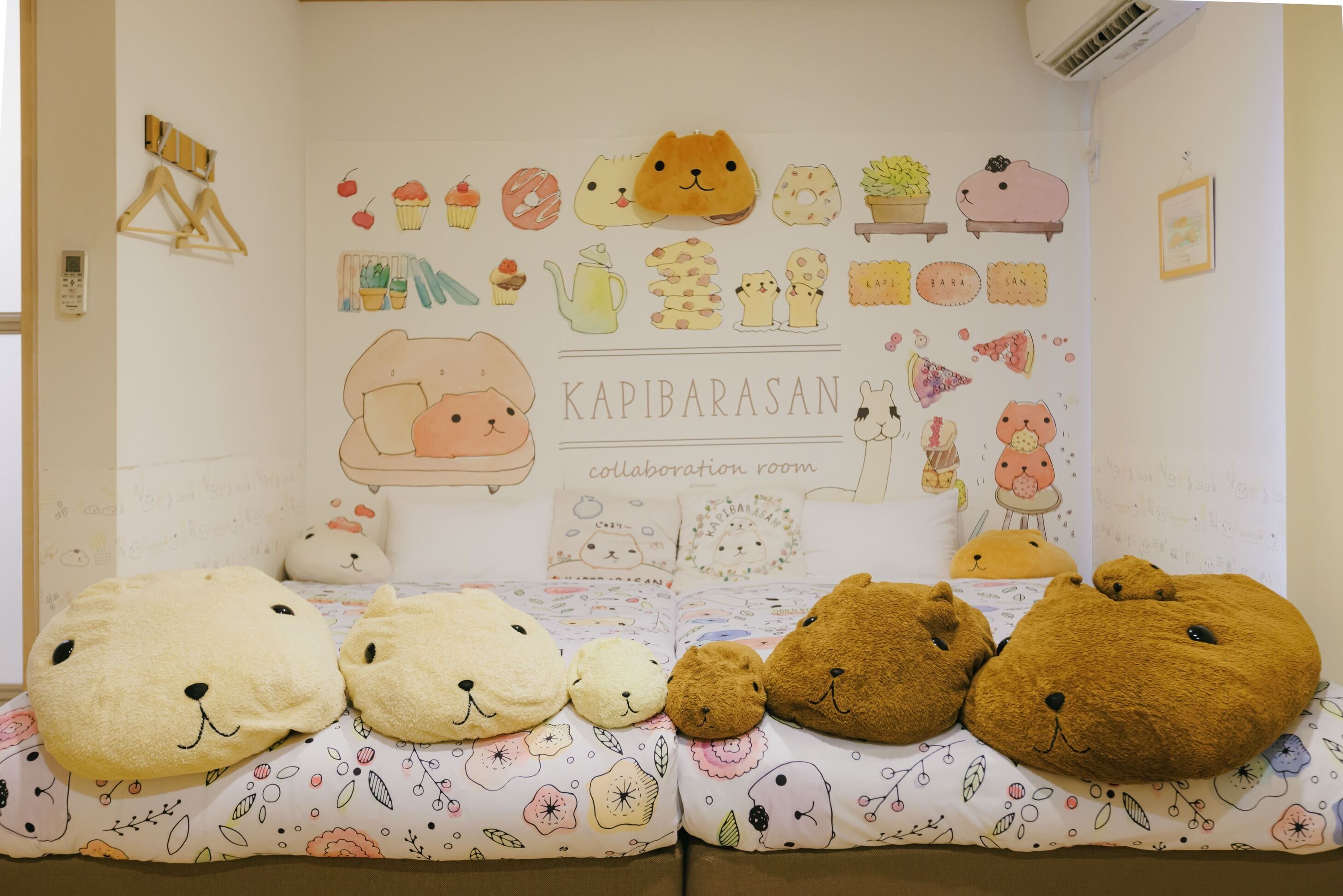 A room with a white wall decorated with various illustrations and a sign that reads 'KAPIBARASAN collaboration room.' There are plush animal pillows on the bed.