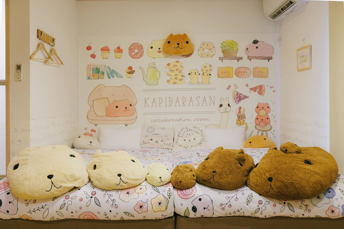 A room with a white wall decorated with various illustrations and a sign that reads 'KAPIBARASAN collaboration room.' There are plush animal pillows on the bed.