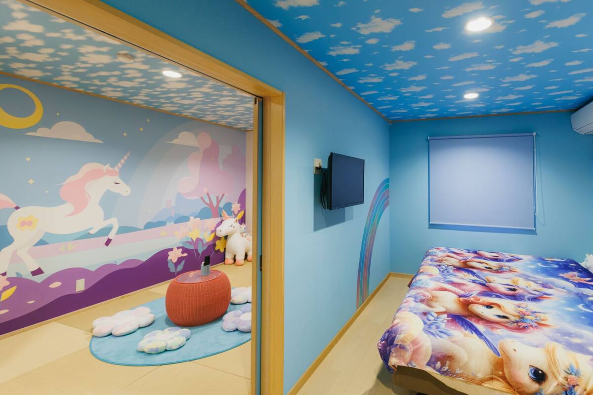 A room with a mural of unicorns, a bed with a unicorn-themed bedspread, a TV, and a ceiling with a cloud design.